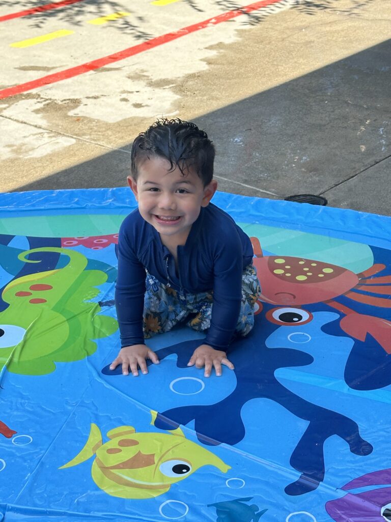A young boy sits on a blue mat decorated with colorful fish, smiling and enjoying his playtime.