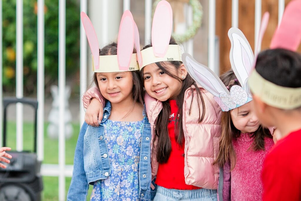 Three children wearing colorful bunny ears joyfully hold hands in a playful outdoor setting.